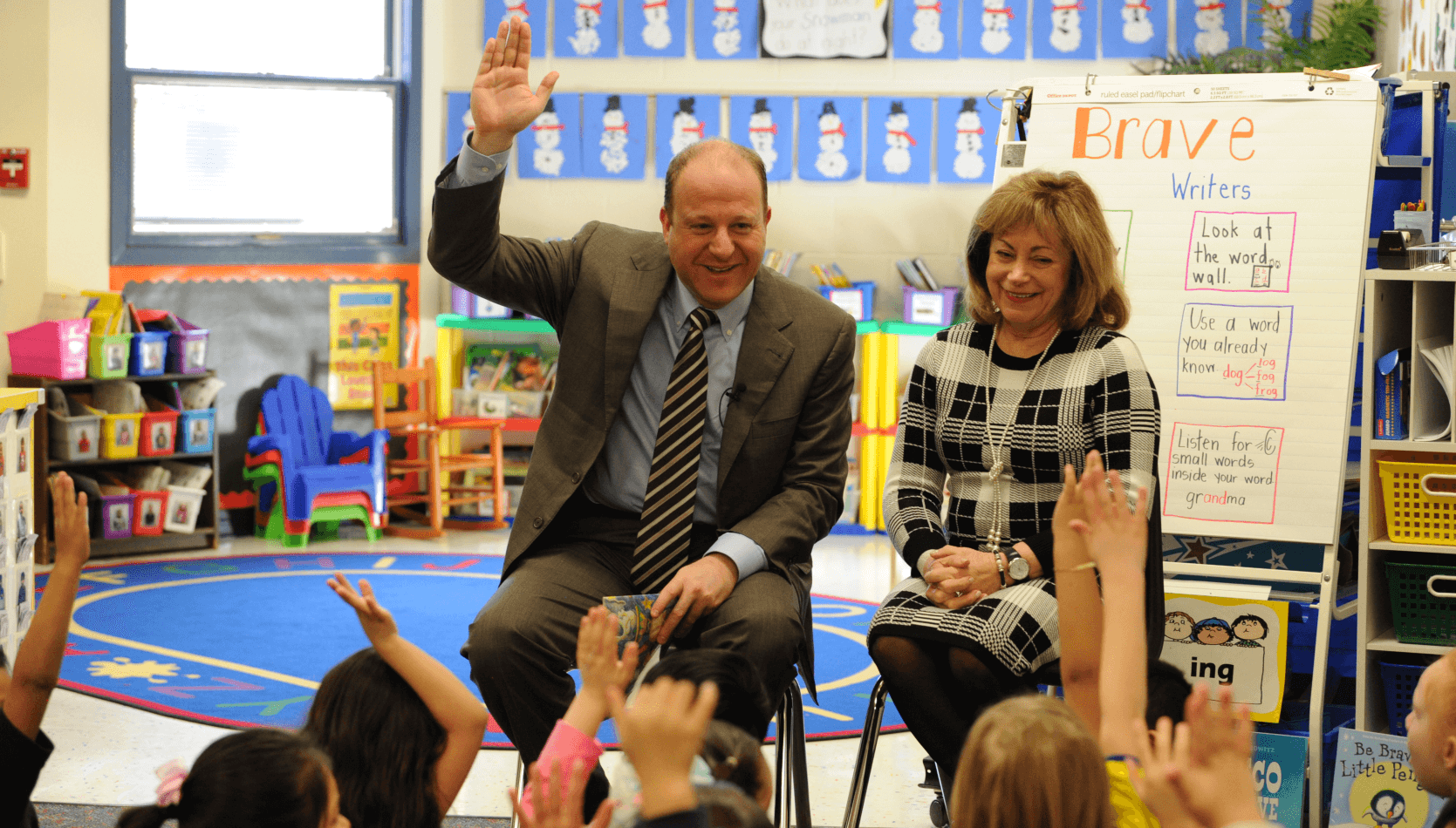 Governor Jared Polis smiles and raises his hand in front of a room of elementary school children. Seated next to him is Lt. Governor Primavera..